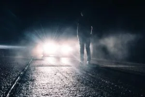 Pedestrian in street with car approaching and headlights illuminating pedestrian.